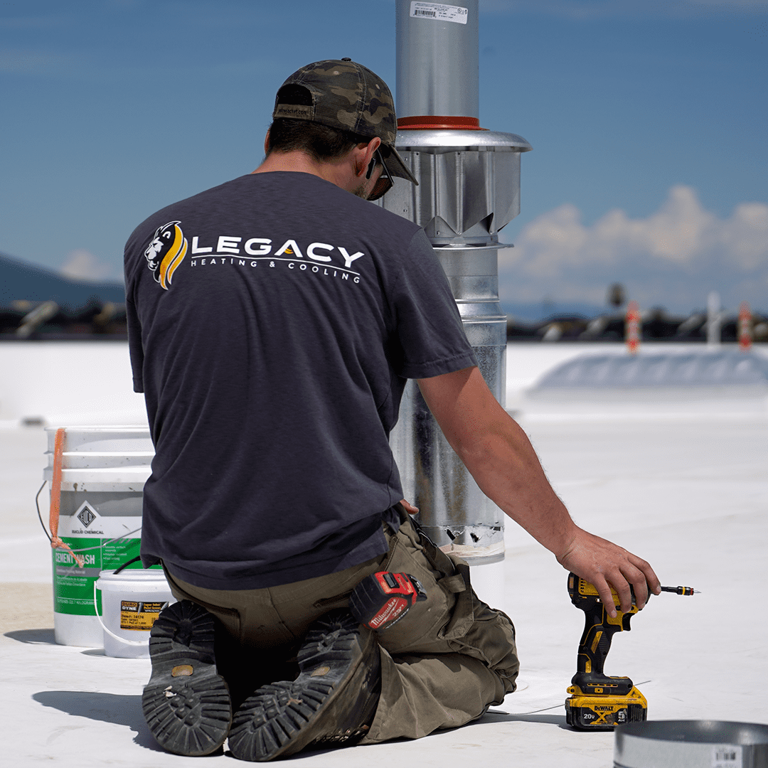 A worker kneels on a flat rooftop next to ventilation equipment, holding a power drill. Sporting a "Legacy Heating & Cooling" shirt and camouflage cap, he represents North Idaho business and small business success in HVAC innovation.