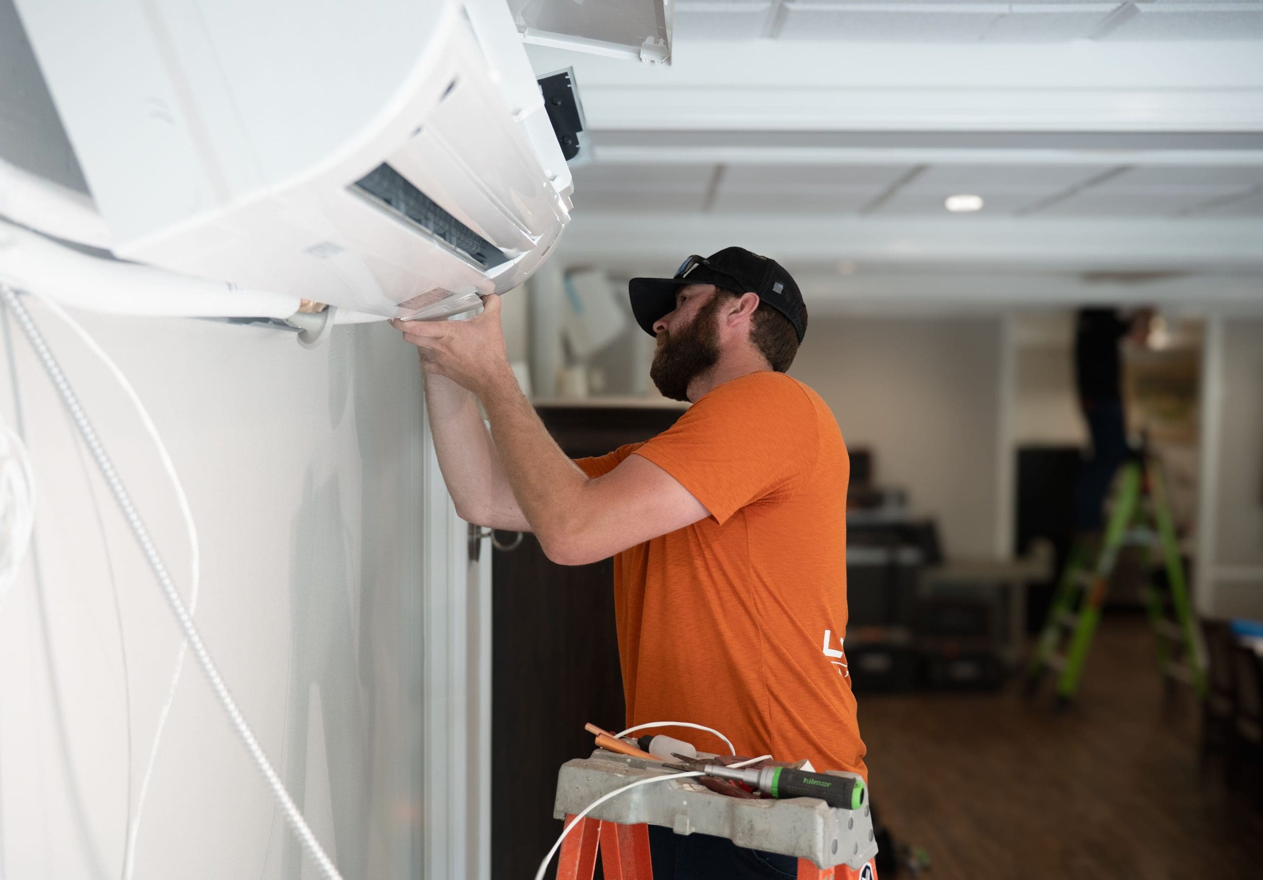 A man in an orange shirt stands on a step stool, installing or repairing a wall-mounted air conditioning unit indoors, demonstrating expertise in commercial cooling solutions.