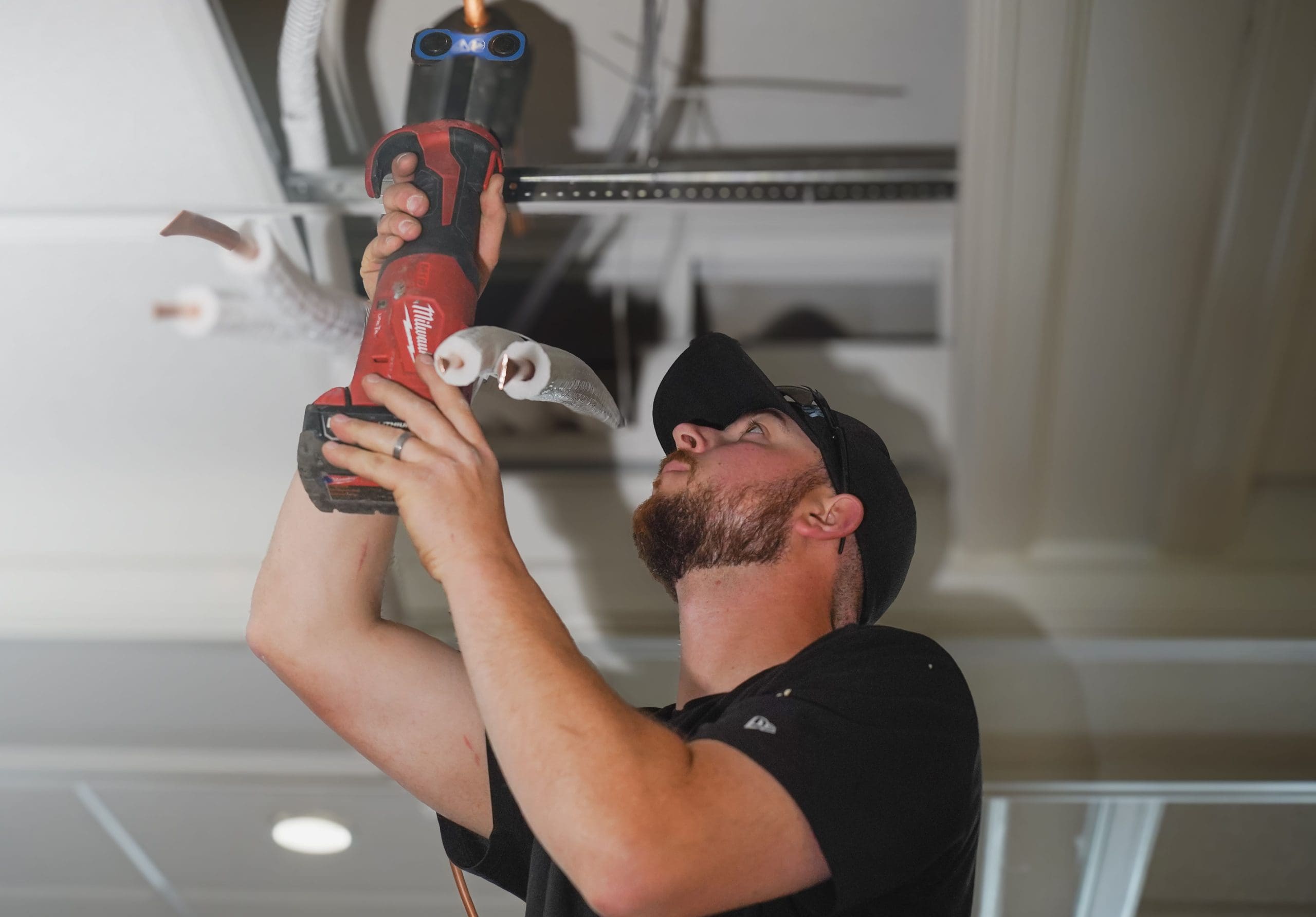 A man wearing a cap uses a power tool to work on electrical wiring in a ceiling, surrounded by exposed cables during a commercial HVAC new construction project.